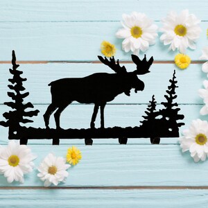 May include: Black metal moose silhouette key holder with two pine tree silhouettes on either side. The key holder is on a light blue wooden background with white daisies.