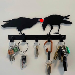 May include: Black metal key rack shaped like two crows with a red circle between them. The rack has five hooks with various keys and keychains hanging from them. The keychains include a green keychain with a small bottle, a black keychain with a small metal object, a brown keychain with a small metal object, a silver keychain with a small metal object, and a silver keychain with a small metal object. The keychains also include a small green keychain with the text "SERVICE KEY" and a small brown keychain with the text "D+D".