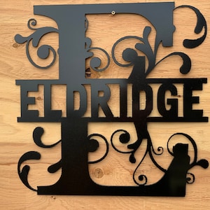 May include: Black metal monogram with the letter "E" and the name "ELDRIDGE" in a decorative font. The letter "E" has a floral design.