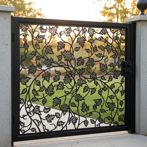 May include: Black metal garden gate with a decorative leaf and vine design. The gate is set between two concrete pillars and has a latch on the right side. The background shows green grass and trees.