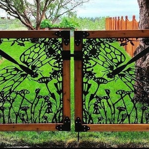 May include: A pair of wooden gates with a black metal design featuring butterflies and flowers. The gates are open and show a green lawn behind them.