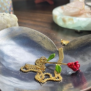 Peut inclure: Un collier en cha&icirc;ne dor&eacute;e avec un pendentif en forme de rose rouge. La rose est en m&eacute;tal et a des feuilles vertes. Le collier est pos&eacute; sur un plat en forme de c&oelig;ur.