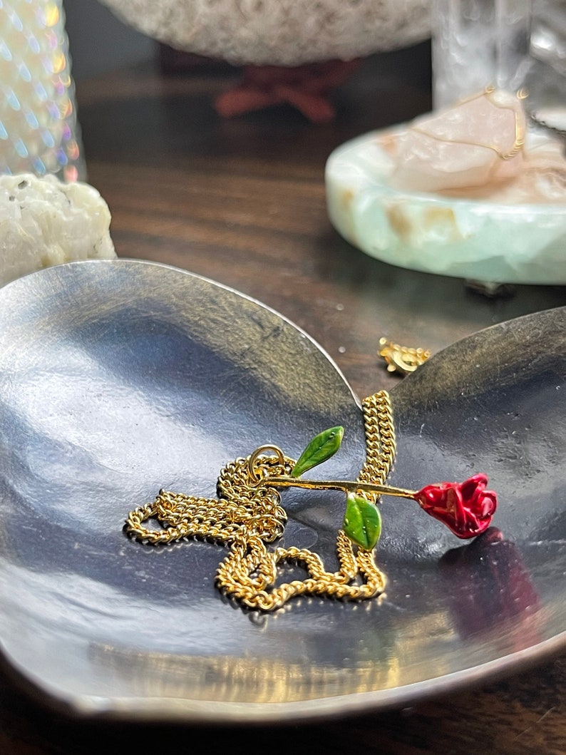 Peut inclure: Un collier en cha&icirc;ne dor&eacute;e avec un pendentif en forme de rose rouge. La rose est en m&eacute;tal et a des feuilles vertes. Le collier est pos&eacute; sur un plat en forme de c&oelig;ur.