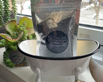 Peut inclure: Une baignoire en porcelaine blanche avec une bordure noire et trois pieds. Un sac en plastique transparent contenant un mélange de sel de bain se trouve à l'intérieur de la baignoire. L'étiquette sur le sac indique "New Moon Soak A Premium Bath Salt Blend with Amethyst Crystal infused with moon magick Net Wt. 150 g (0.1) Lavender/Lemongrass".