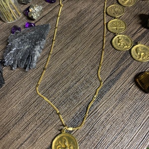 May include: Gold chain necklace with a gold pendant featuring a zodiac symbol. The pendant is oval shaped and has a detailed design of a mythical creature.