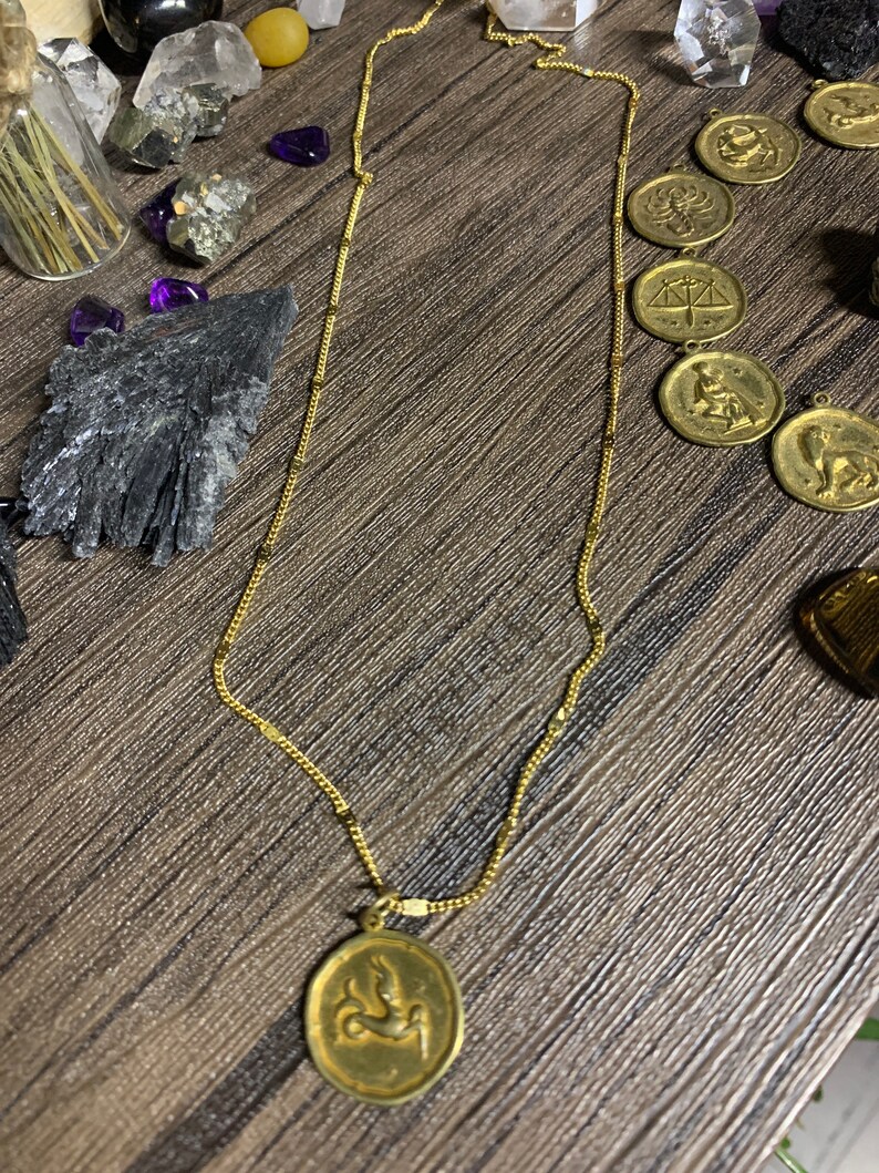 May include: Gold chain necklace with a gold pendant featuring a zodiac symbol. The pendant is oval shaped and has a detailed design of a mythical creature.