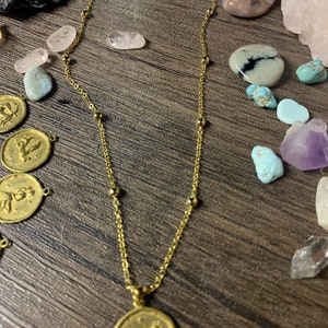 May include: A gold chain necklace with a gold pendant featuring a zodiac symbol. The pendant is round and has a detailed design of a bull. The necklace is on a wooden surface with various crystals and stones.