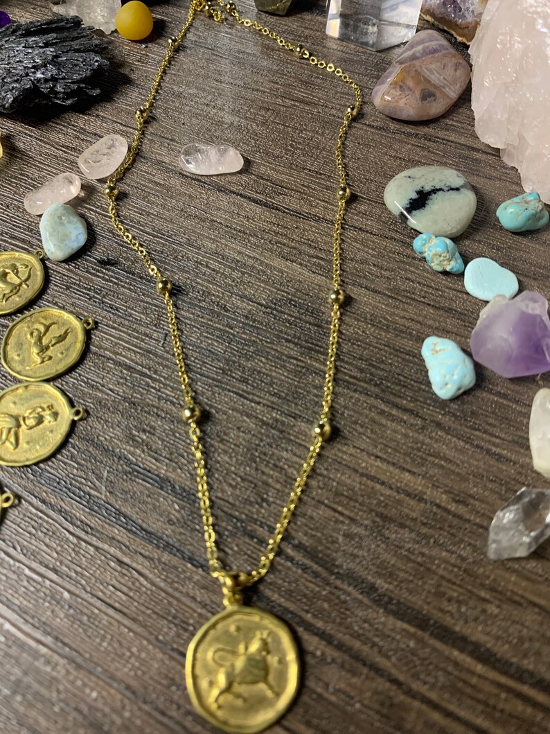 May include: A gold chain necklace with a gold pendant featuring a zodiac symbol. The pendant is round and has a detailed design of a bull. The necklace is on a wooden surface with various crystals and stones.
