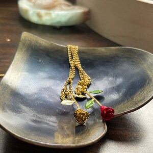 Peut inclure: Un collier en cha&icirc;ne dor&eacute;e avec deux pendentifs en forme de rose, l'un dor&eacute; et l'autre rouge, sur un plat en forme de c&oelig;ur gris fonc&eacute;.