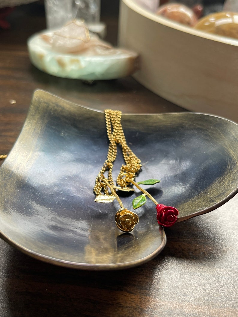 Peut inclure: Un collier en cha&icirc;ne dor&eacute;e avec deux pendentifs en forme de rose, l'un dor&eacute; et l'autre rouge, sur un plat en forme de c&oelig;ur gris fonc&eacute;.