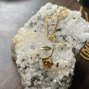 Peut inclure: Un collier avec un pendentif en forme de rose dor&eacute;e sur une cha&icirc;ne d&eacute;licate. La rose est orn&eacute;e de feuilles et repose sur un lit de cristaux blancs.