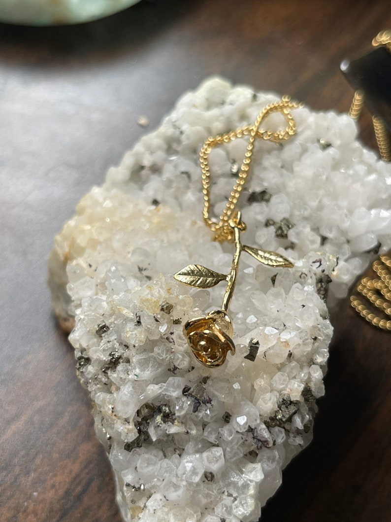 Peut inclure: Un collier avec un pendentif en forme de rose dor&eacute;e sur une cha&icirc;ne d&eacute;licate. La rose est orn&eacute;e de feuilles et repose sur un lit de cristaux blancs.