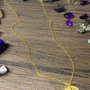 May include: A gold chain necklace with a round pendant featuring a stylized image of a horse. The pendant is on a wooden surface with various crystals and stones.