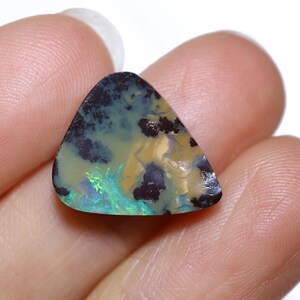 Puede incluir: Una piedra preciosa de &oacute;palo triangular con una mezcla de colores. La piedra presenta manchas marrones oscuras, parches amarillos y azules y un destello verde. El &oacute;palo se sostiene entre dos dedos.