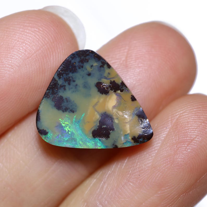 Puede incluir: Una piedra preciosa de &oacute;palo triangular con una mezcla de colores. La piedra presenta manchas marrones oscuras, parches amarillos y azules y un destello verde. El &oacute;palo se sostiene entre dos dedos.