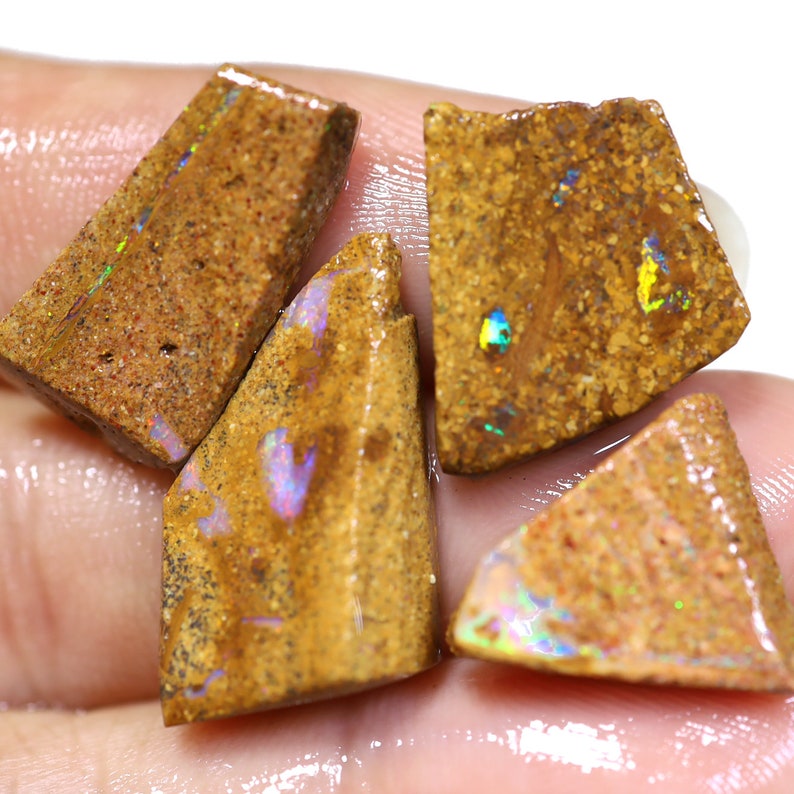 May include: Four rough-cut opal stones with a brown matrix and iridescent flashes of color. The opals have a rough, natural texture and display a range of colors, including blue, green, and yellow. The stones are irregularly shaped and appear to be freshly cut.