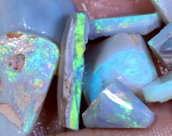 Puede incluir: Una colección de piedras preciosas de ópalo en bruto e iridiscente de varias formas y tamaños. Los ópalos tienen un color azul, verde y blanco con un brillo arcoíris.