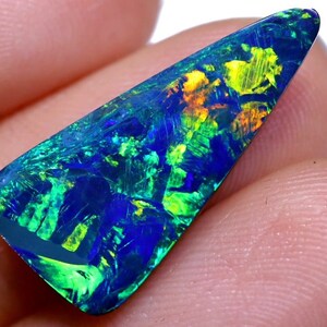 May include: A dark blue, green, and yellow opal gemstone with a triangular shape. The opal is iridescent and has a variety of colors.