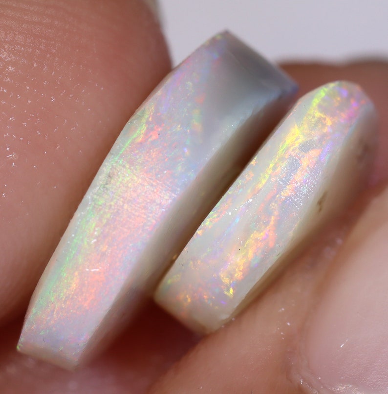 Op de afbeelding: Twee witte opaal cabochons met iriserende regenboogflitsen. De opalen zijn gesneden in een afgeronde rechthoekige vorm.