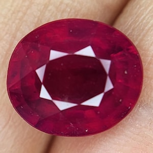 May include: A deep red ruby gemstone with a faceted cut. The gemstone has a star-shaped pattern in the centre.