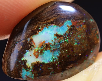 Puede incluir: Un cabujón de ópalo marrón oscuro con un patrón iridiscente azul verdoso y blanco. El ópalo tiene una textura rugosa y natural.