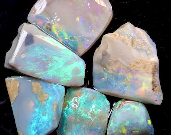 Puede incluir: Una colección de siete piedras de ópalo en bruto con colores iridiscentes azules, verdes y blancos. Las piedras son de varias formas y tamaños.