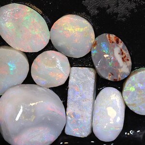 Op de afbeelding: Een verzameling witte opaal edelstenen met iriserende regenboogkleuren. De opalen zijn in verschillende vormen en maten, waaronder ovaal, rond en rechthoekig.