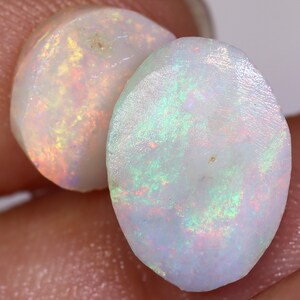 Op de afbeelding: Twee ovale, witte opalen met iriserende regenboogkleuren. De opalen zijn doorschijnend en hebben een glad, gepolijst oppervlak.