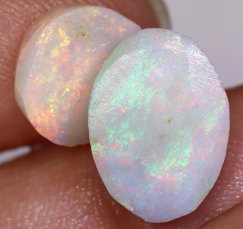 Op de afbeelding: Twee ovale, witte opalen met iriserende regenboogkleuren. De opalen zijn doorschijnend en hebben een glad, gepolijst oppervlak.