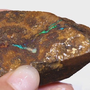 Puede incluir: Un espécimen de ópalo en bruto con una matriz marrón y destellos iridiscentes verdes y azules. La gema muestra una superficie natural sin pulir, mostrando su belleza en bruto. Los colores del ópalo brillan en su superficie.
