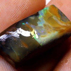 May include: A dark, iridescent opal with flashes of green, blue, and gold. The opal is a rectangular shape and has a rough, natural finish.
