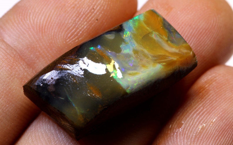 May include: A dark, iridescent opal with flashes of green, blue, and gold. The opal is a rectangular shape and has a rough, natural finish.