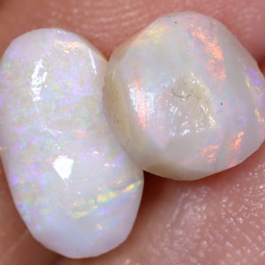 Op de afbeelding: Twee witte opaal edelstenen met iriserende regenboogflitsen. De opalen zijn in verschillende vormen geslepen, een is ovaal en de andere is rond.