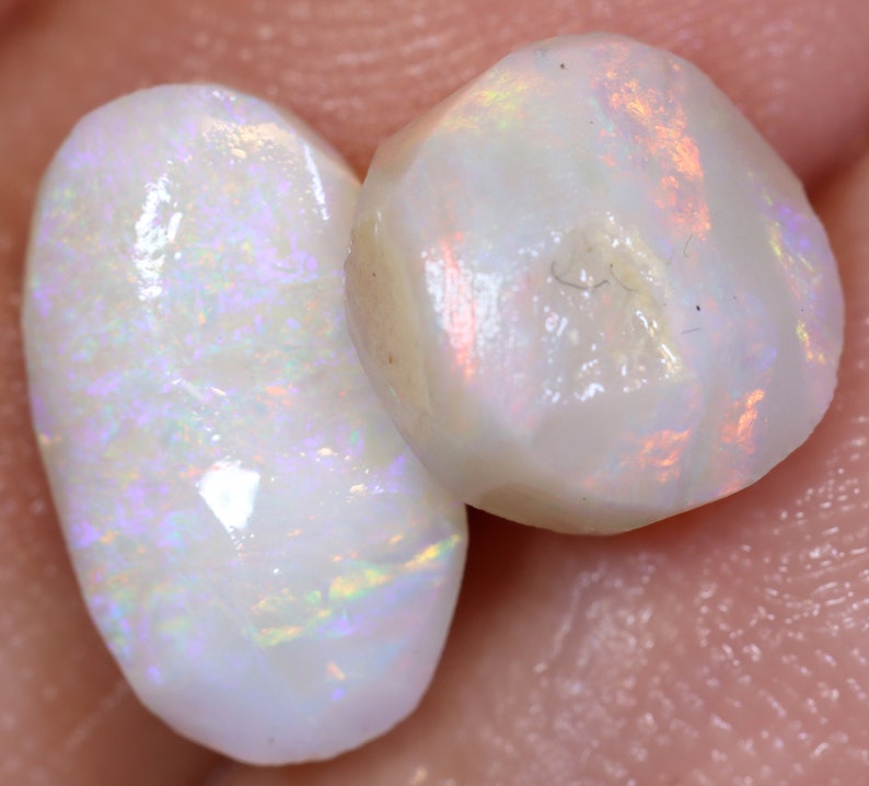 Op de afbeelding: Twee witte opaal edelstenen met iriserende regenboogflitsen. De opalen zijn in verschillende vormen geslepen, een is ovaal en de andere is rond.