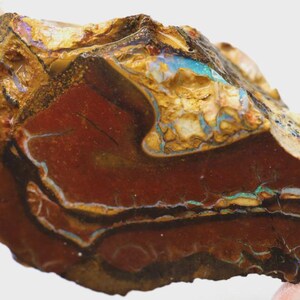 Könnte beinhalten: Ein roher Opalstein mit einer Mischung aus Farben. Der Stein hat eine tiefrote Basis mit Schichten aus Braun, Gold und blaugrünen Farbtönen. Die Oberfläche hat ein strukturiertes Aussehen mit natürlichen Mustern und Rissen.