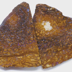 Könnte beinhalten: Zwei Teile eines polierten Opal-Edelsteins, die ein Mosaikmuster in Braun-, Gold- und Lilatönen aufweisen. Der Opal hat eine einzigartige Textur und eine natürliche Form, mit einem weißen Fleck in der Mitte eines Teils.