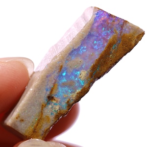 Puede incluir: Una gema de ópalo en bruto con una vibrante exhibición de colores. La piedra presenta una mezcla de tonos azules, morados y verdes dentro de una matriz marrón y beige. La superficie del ópalo tiene una textura natural e irregular.