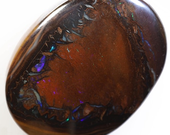 Puede incluir: Una piedra preciosa de ópalo marrón, negro e iridiscente con una forma ovalada lisa. El ópalo tiene una base marrón oscura con destellos de azul, verde y morado.
