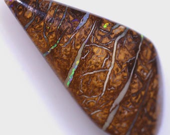 Puede incluir: Una piedra preciosa pulida en forma de lágrima, con un color base marrón y un patrón complejo de vetas blancas y marrón oscuro. Destellos iridiscentes de verde, azul y púrpura son visibles dentro de la piedra, agregando un contraste vibrante a los tonos terrosos.