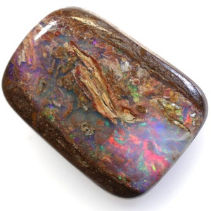Puede incluir: Un gran ópalo rectangular marrón con colores iridiscentes de púrpura, verde, rojo y azul. El ópalo tiene una textura natural y rugosa y está engastado en una matriz marrón oscuro.