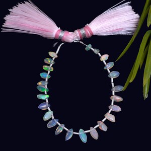 May include: A necklace of opal gemstones strung on silver wire, with a pink tassel at the top. The oval-shaped opals show iridescent colours of green, blue, and pink. The background is black.