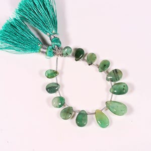 Op de afbeelding: Een streng traanvormige groene smaragd kralen, geregen op een zilveren draad. De kralen variëren in tint van licht- tot donkergroen. Een turquoise kwast is bevestigd aan de bovenkant van de streng. De kralen worden gepresenteerd tegen een witte achtergrond.
