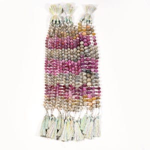 Puede incluir: Cadenas de cuentas de piedras preciosas en tonos rosa, gris y amarillo, con una borla en cada extremo. Las cuentas son de varios tamaños y formas. Las borlas son blancas y verde pastel.