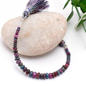 May include: A necklace of multi-coloured gemstone beads, including shades of red, blue, and purple. The beads are strung together and attached to a tassel with silver-coloured metal accents. The tassel features strands of purple, blue, and green threads.