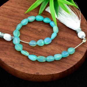 May include: A strand of oval-shaped, light blue opal beads. The beads are strung on a silver wire and are displayed on a wooden surface. A white tassel with silver accents is visible in the background. The beads have a subtle iridescent quality.