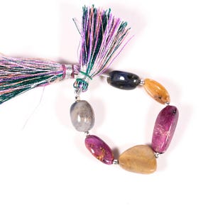 Puede incluir: Un collar de cuentas de piedras preciosas multicolores en tonos morado, amarillo y azul, conectadas por cuentas de metal plateado. Una borla con hilos morados, verdes y blancos está adjunta. Las cuentas tienen forma irregular.