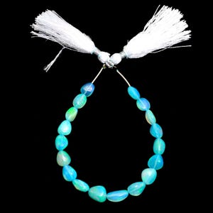 Puede incluir: Un collar de cuentas de ópalo ovaladas y pulidas en tonos azules y verdes. Las cuentas están ensartadas con una borla blanca en cada extremo, formando un bucle. El fondo es negro.