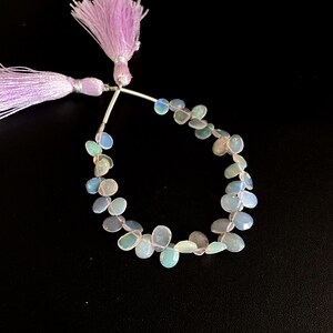 Puede incluir: Un collar de cuentas de ópalo iridiscente en forma de lágrima. Las cuentas presentan tonos azul pálido, verde y rosa. El collar está unido a dos borlas lavanda con detalles plateados, sobre un fondo negro.
