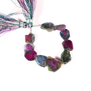 May include: A strand of raw ruby gemstone beads in shades of deep red, blue, and green. The beads are strung together with a silver-colored wire and attached to a tassel with green, purple, and white threads.