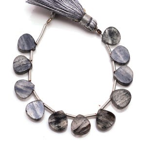May include: A strand of heart-shaped, gray-blue gemstone beads with a silver-colored wire and a gray tassel. The beads have a marbled appearance with darker gray and black inclusions. The tassel is made of gray threads.